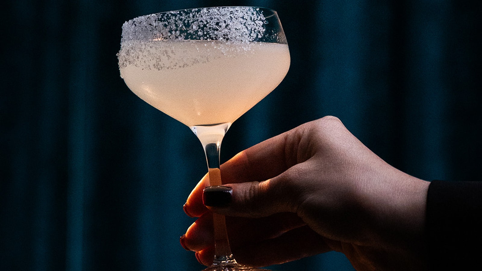 Hand met een glas cocktail met gezouten rand tegen een blauw fluwelen gordijn